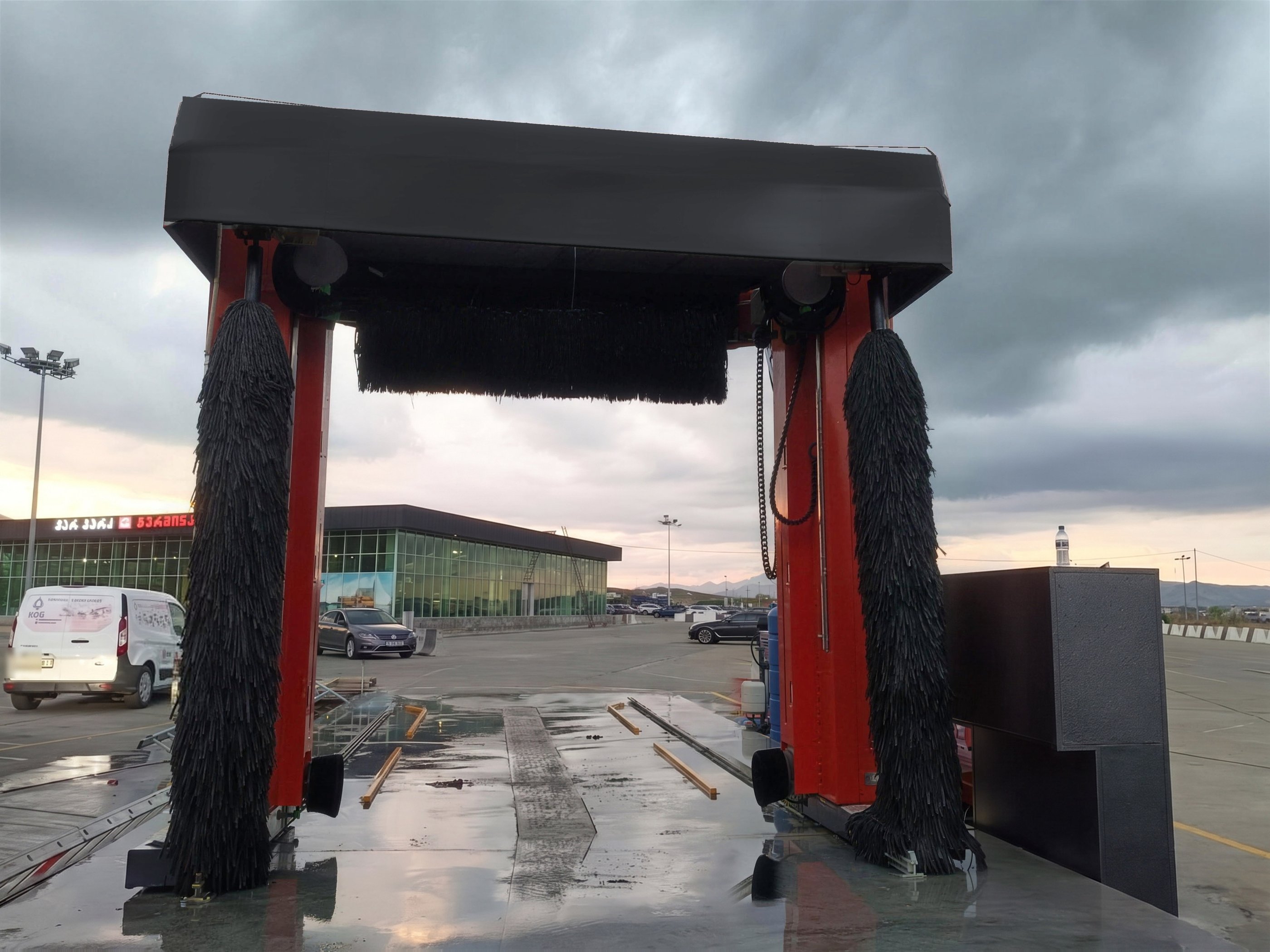 A red KKE X1.1 rollover car wash seen low and close from inside its bay, both vertical brushes hanging from the portal either side of the horizontal roof brush, under a storm sky