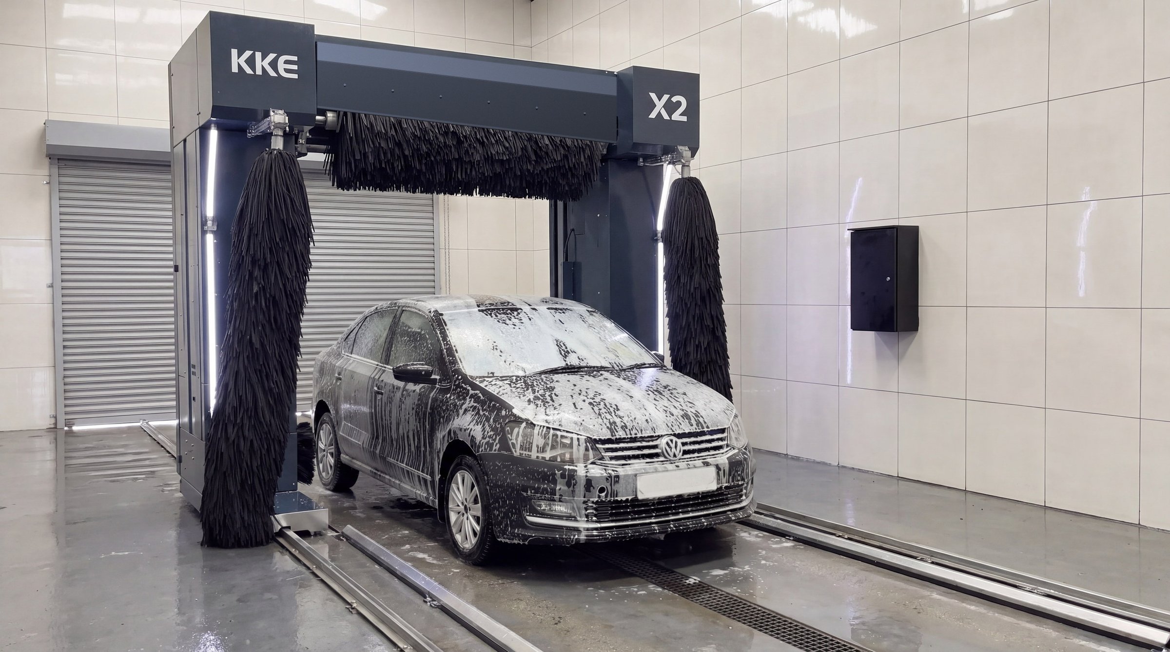 A saloon car sheeted head to tail in white foam standing between the brushes of a KKE X2, the bay's roller shutter closed behind it