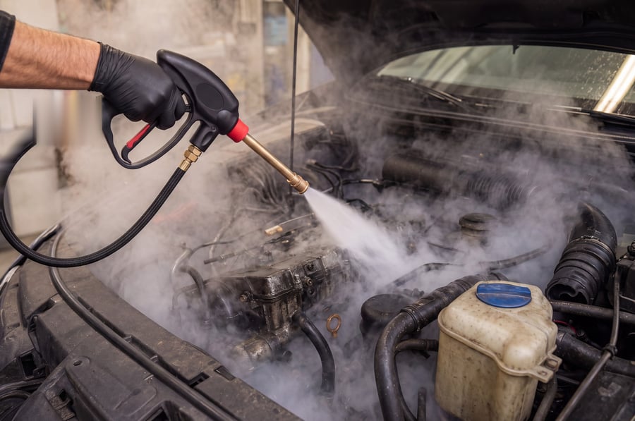 A technician in black gloves steam cleaning a dirty engine with a steam gun and brass lance, the bonnet propped open and a cloud of steam rolling through the bay off the cam cover