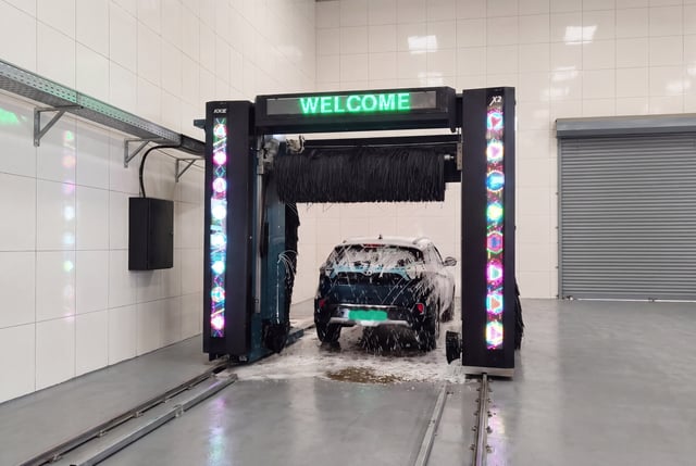 A KKE X2 rollover standing over a car in a tiled bay, WELCOME lit on the programme display
