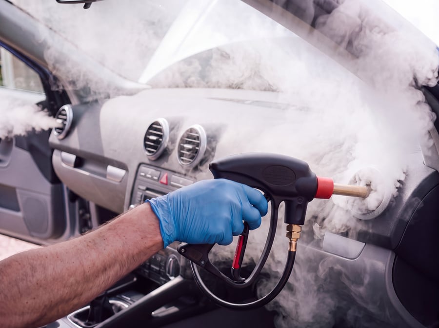 A gloved hand steam cleaning a car's dashboard air vents with a lance, steam coming back out through the vent blades