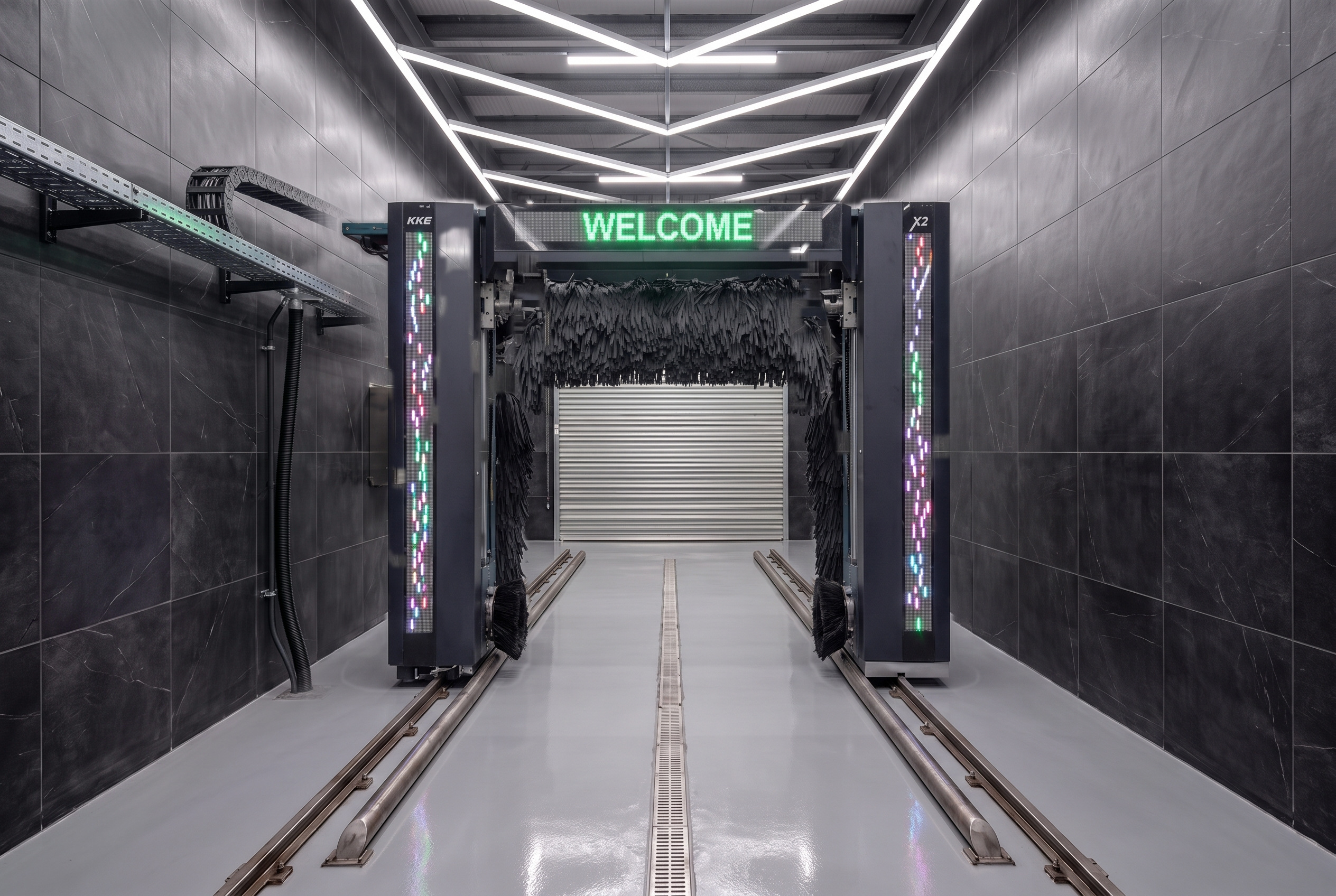 A KKE X2 automatic car wash gantry parked at the far end of a black-tiled wash bay, its two LED columns lit and WELCOME showing green on the beam, with the bay's floor rails running toward the camera down an empty grey floor