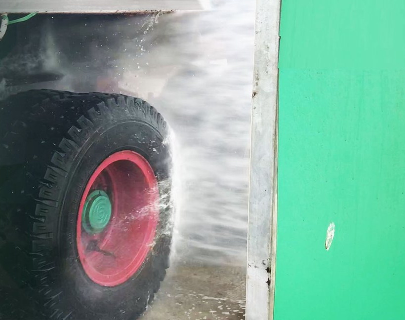A jet of water from a KKE wheel blaster striking the tyre and red wheel rim of a truck, spray bursting off the tread, with the green panel of the machine standing alongside