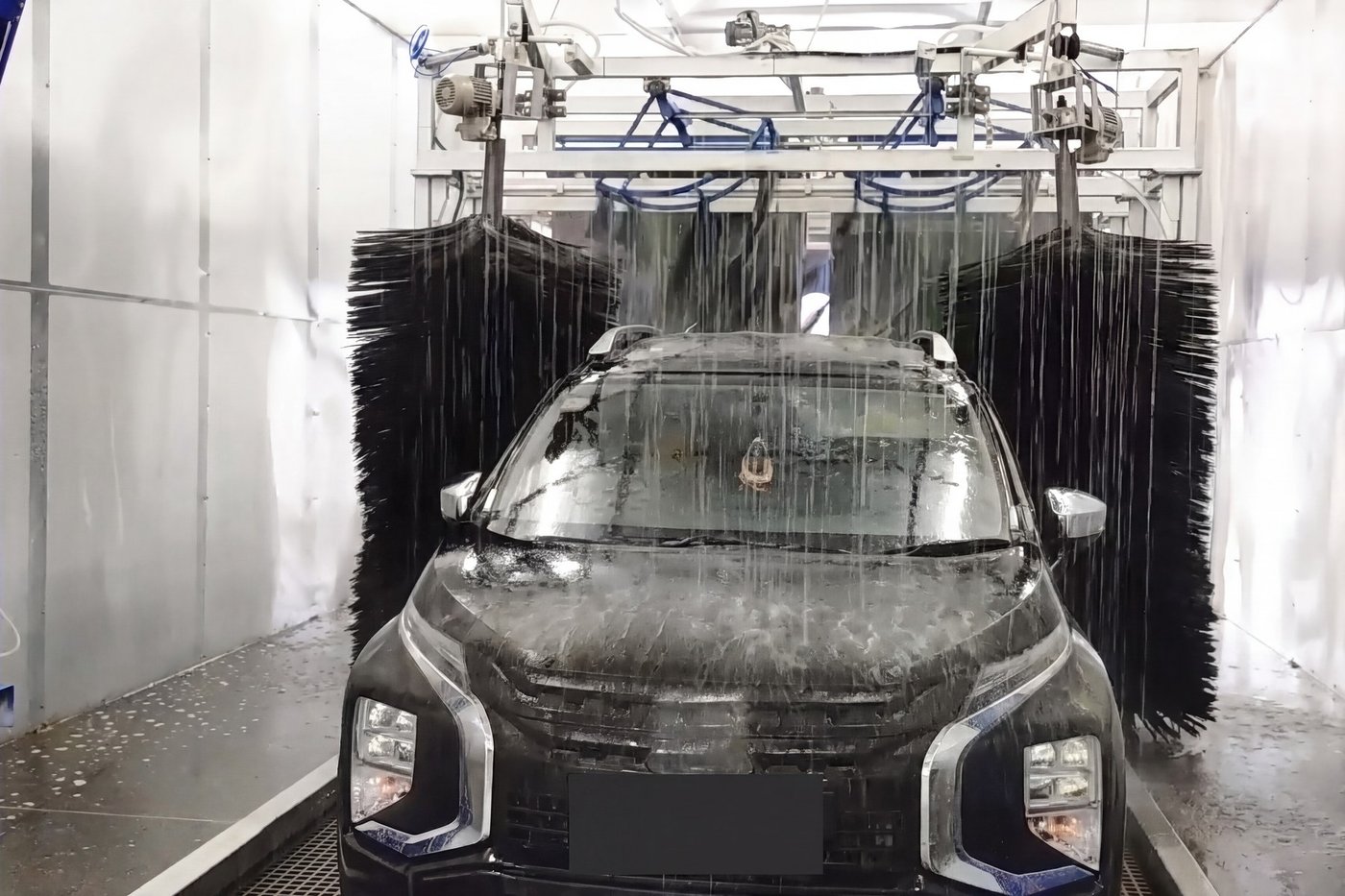 A car head-on between the two vertical wrap brushes of a KKE conveyor tunnel, a full-width sheet of water falling from the gantry above it