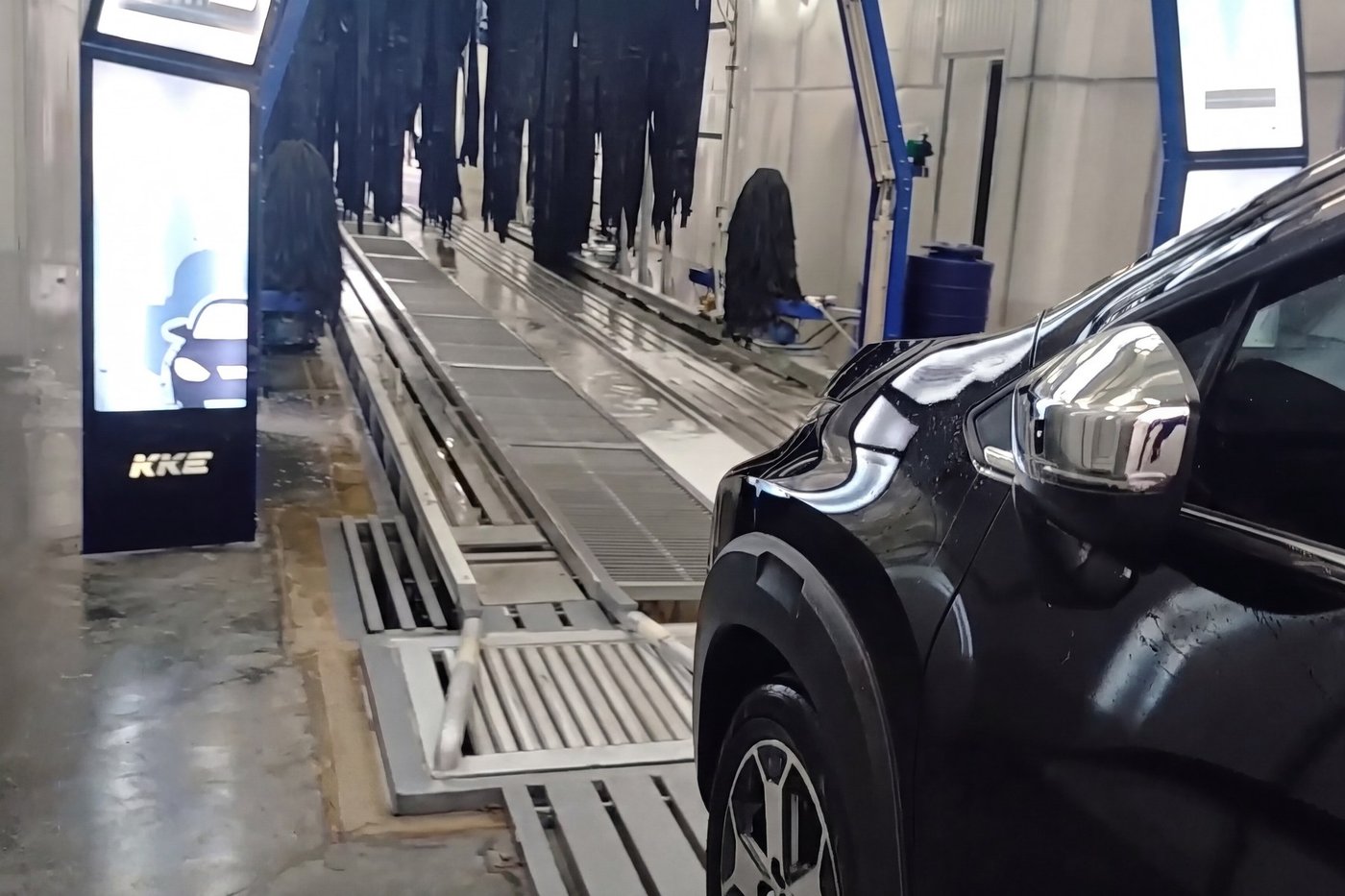 A car standing on the conveyor at the mouth of a KKE tunnel car wash, the guide rails and grating of the conveyor lane running away down the frame past a lit KKE signage totem
