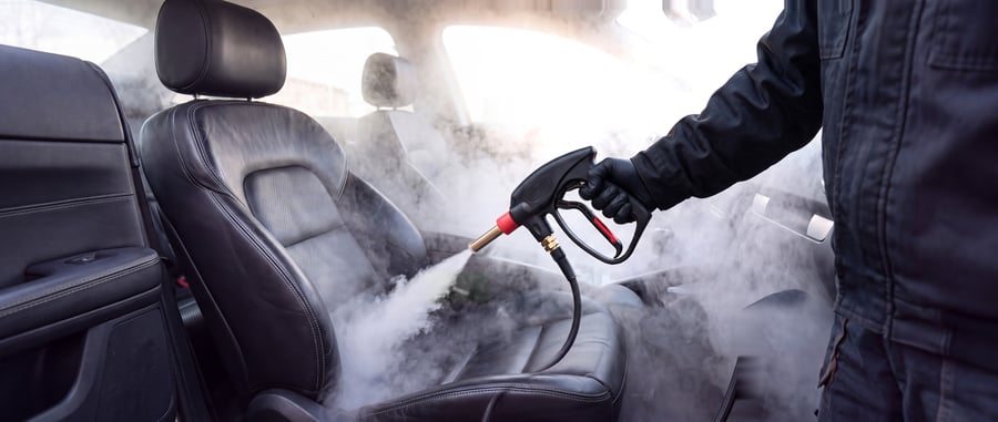 A technician in dark workwear steam cleaning a black perforated leather car seat, a steam gun with a brass lance held over the cushion, steam filling the cabin and the leather armrest in the foreground