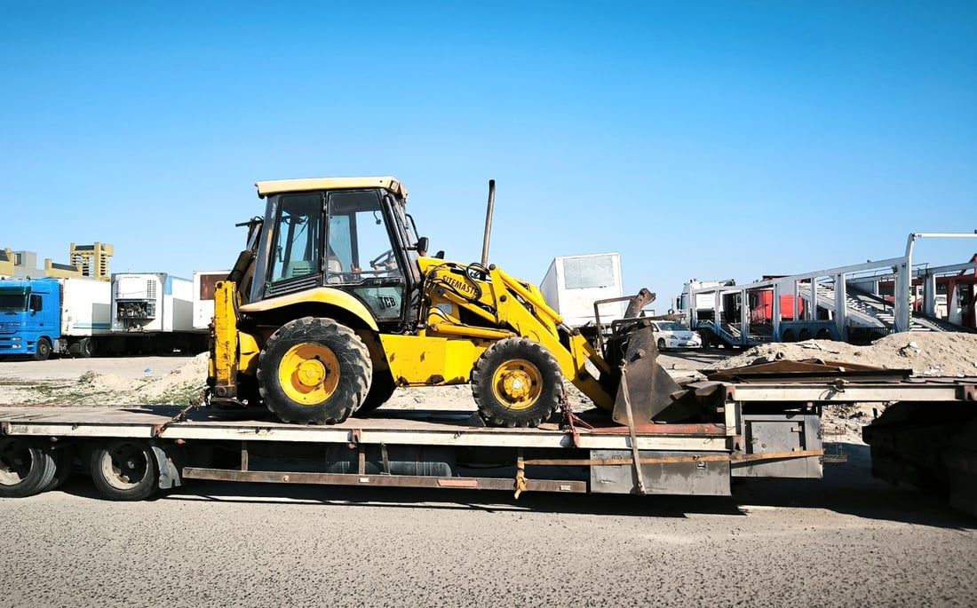 A backhoe loader standing on a raised steel deck with the access ramp folded down behind it