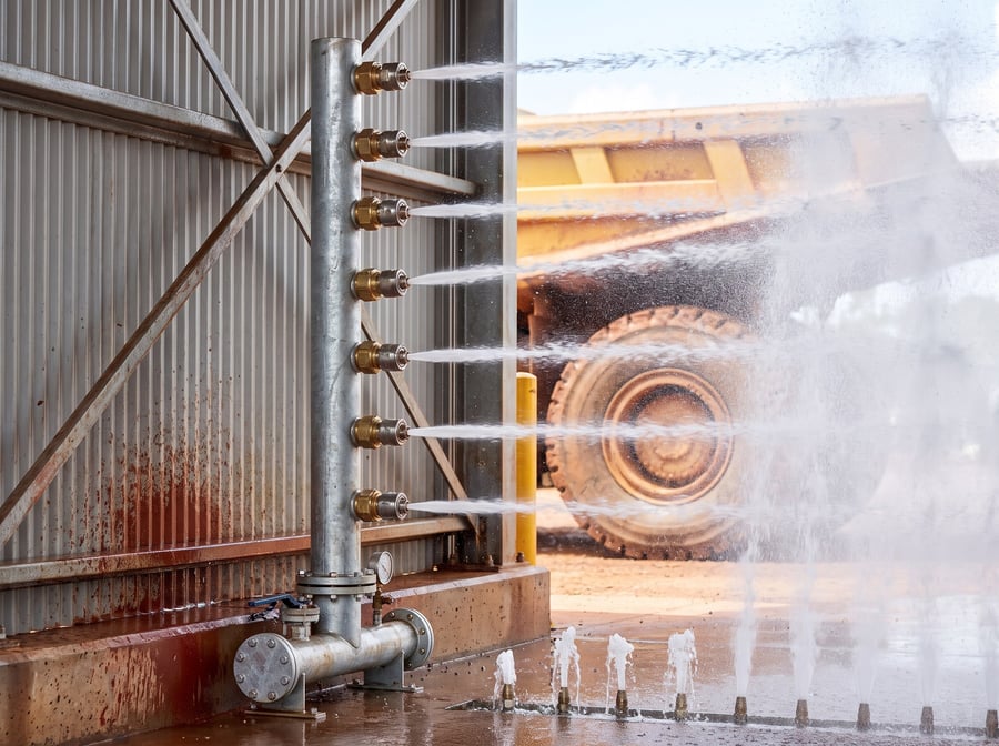 Close view of a full-height vertical nozzle bank firing horizontally inward, with the under-chassis nozzle row in the floor channel throwing water upward and a haul truck wheel beyond