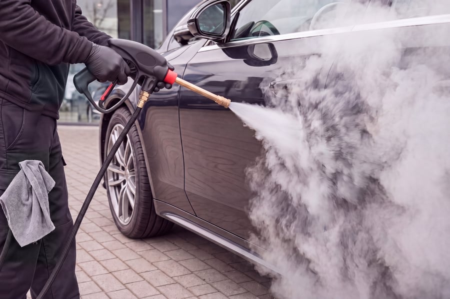 A technician washing the side of a dark car with a steam gun and brass lance on a paved forecourt, a heavy plume of steam flaring off the door and rolling down the sill, the ground under the car dry