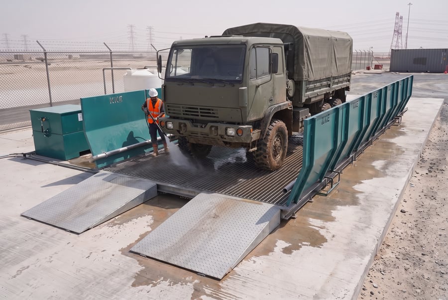 A mud-caked olive military cargo truck standing on a KKE modular wash rack, an operator in hi-vis washing its front wheel with a high-pressure lance