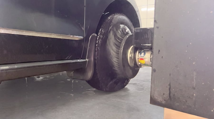 The round black wheel disc brush of a KKE X2 swung out at floor level against a car's wheel, fan-shaped jets of water firing from a nozzle beside it