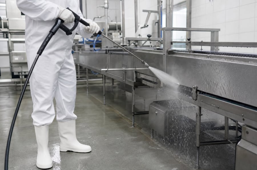 A worker in white hygiene overalls washing down stainless steel food processing equipment and a conveyor with a high pressure lance in a bright clean food factory