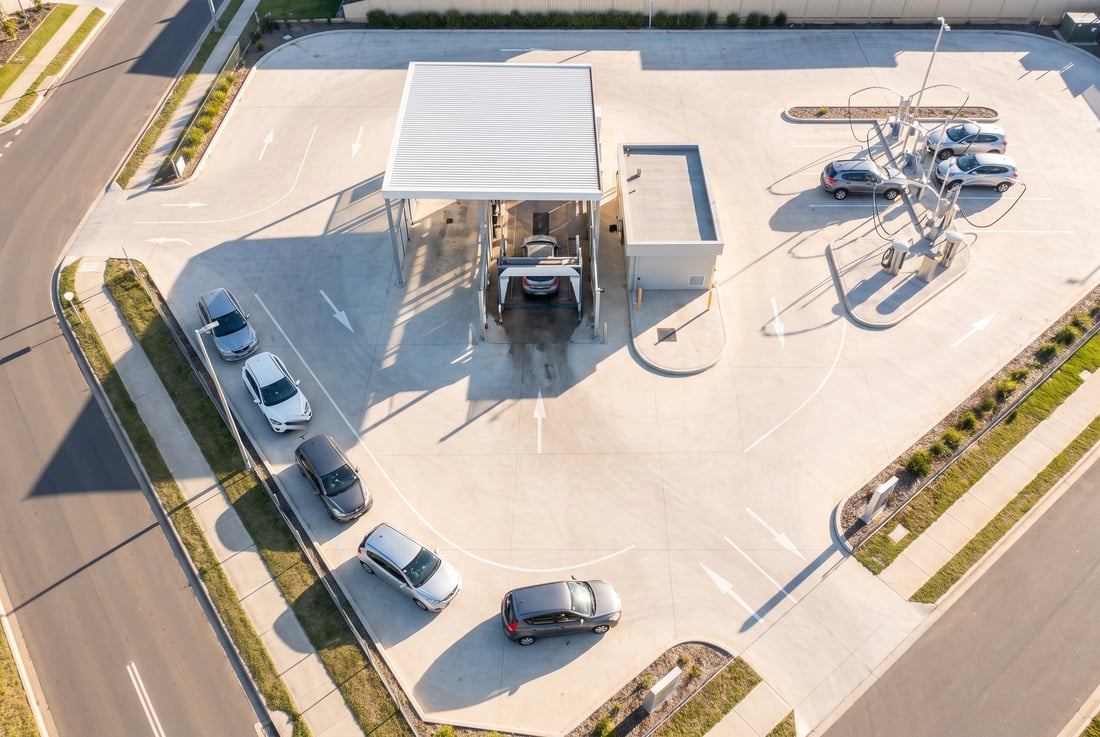 Aerial view of an automatic car wash site: an entry lane with cars waiting, a covered wash bay in the middle of the plot, a separate exit lane and a row of self-service vacuum bays alongside.