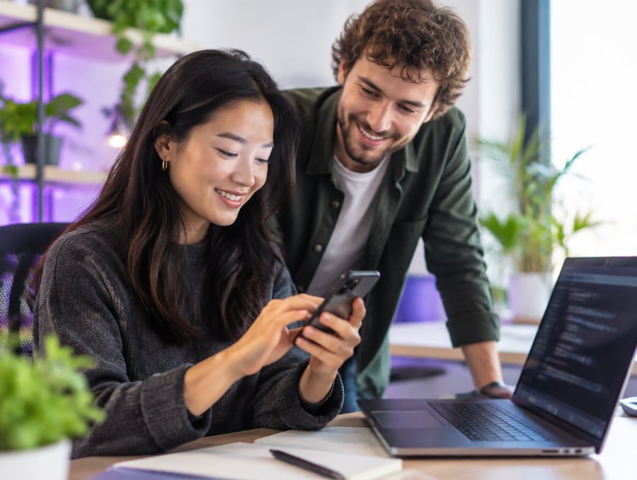 A mobile app developer tests an app on her phone while a colleague looks on, laptop with code beside them