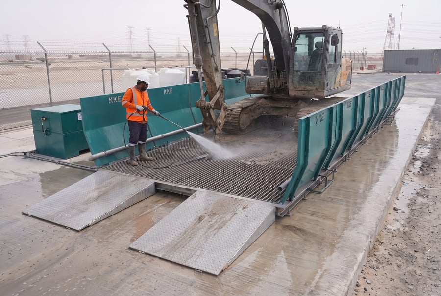 An operator pressure-washing a tracked excavator on a KKE portable wash rack, the splash guard walls catching the spray