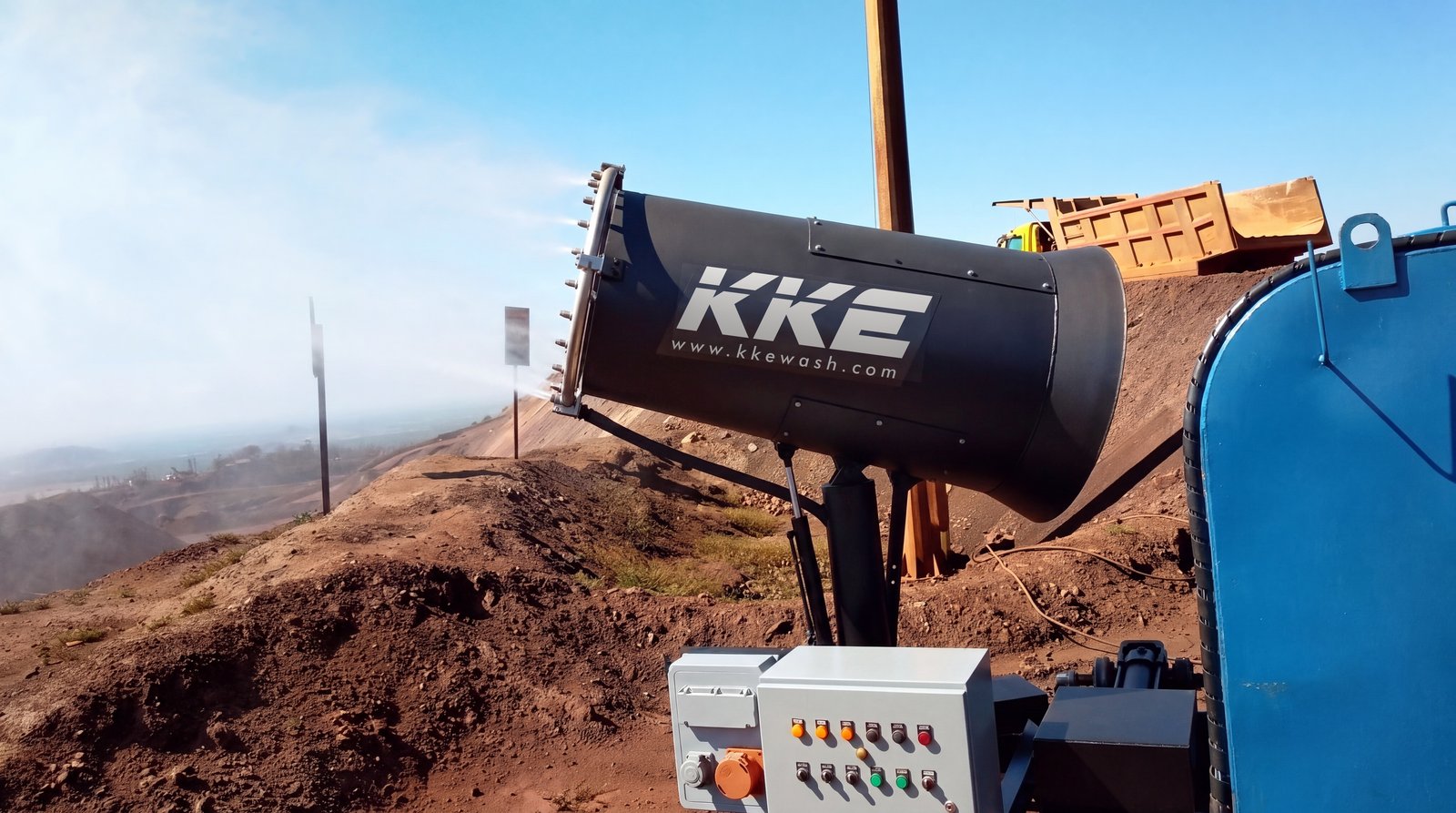 A KKE MistBeam fog cannon misting from an iron-ore bench, open-cast workings in the hazy valley beyond it and a haul truck tipping on the crest behind