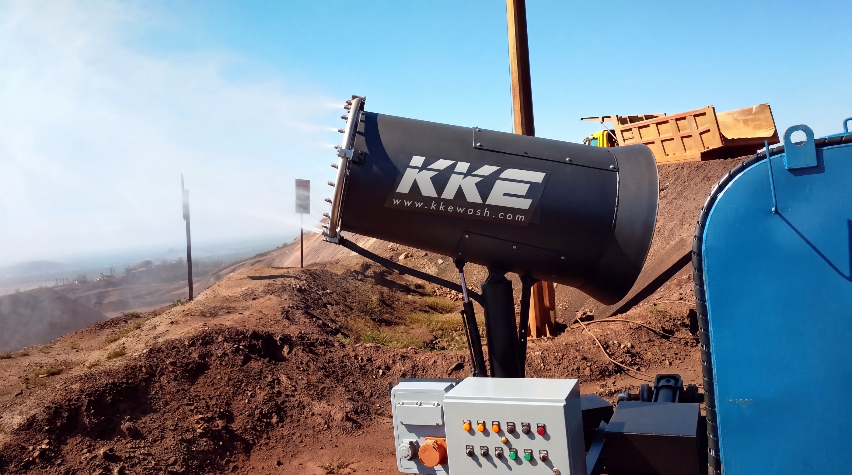 A KKE MistBeam fog cannon misting from an iron-ore bench above a hazy valley, open-cast workings beyond and a haul truck tipping on the crest behind
