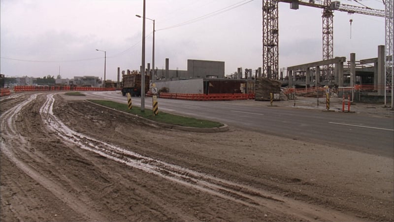 Deep muddy tracks running from churned ground onto clean public asphalt at a construction site exit, tower cranes behind