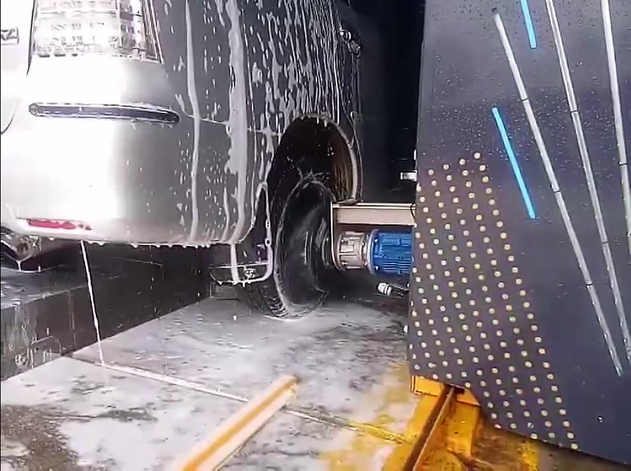 The disc wheel washer of a KKE X1.1 working at floor level against the rear wheel of a foam-covered MPV, its blue geared motor on the slide behind the disc and the column's LED panel alongside