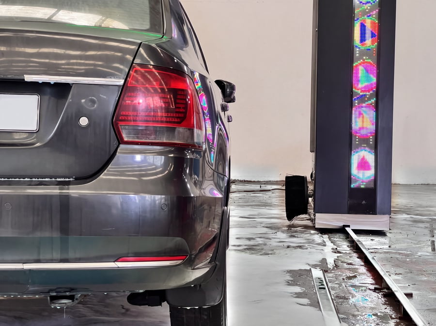 A column LED strip close up, its colours reflected in the dark flank of the car beside it, with the wheel disc brush at its foot