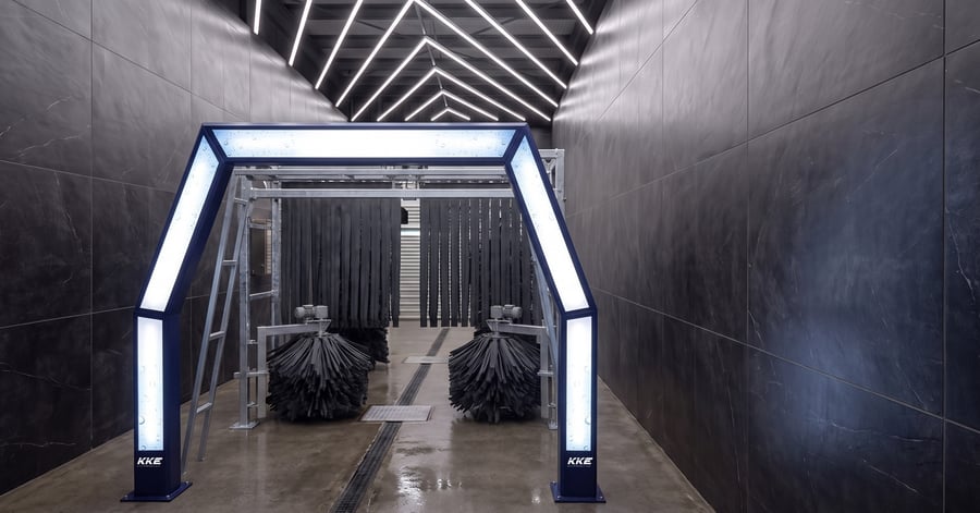 A KKE Z10 fleet car wash standing in a black-tiled wash bay: a navy entry portal with backlit white panels down each leg and across its header, two low charcoal skirt brush drums on the wet floor either side of a covered conveyor slot, and ranks of dark hanging curtain strips running away down the tunnel behind them