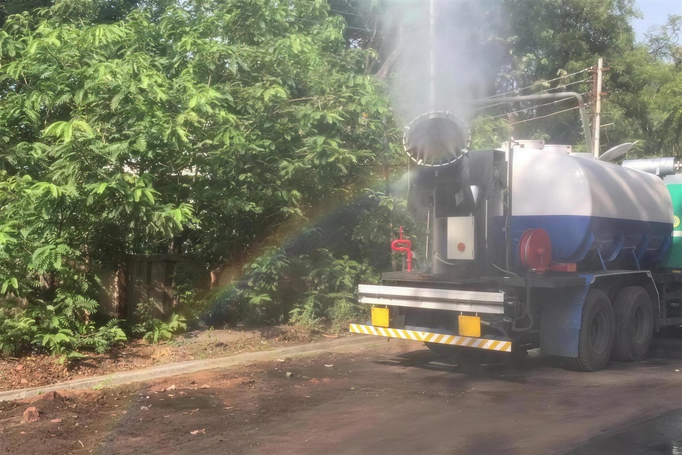 Truck-mounted KKE MistBeam fog cannon misting roadside vegetation from a water tanker, a rainbow forming in the plume