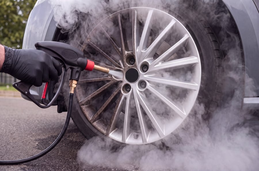 A close-up of a gloved hand holding a steam gun with a brass lance to a silver multi-spoke alloy wheel, a thick cloud of steam boiling out through the spokes and rolling out of the wheel arch