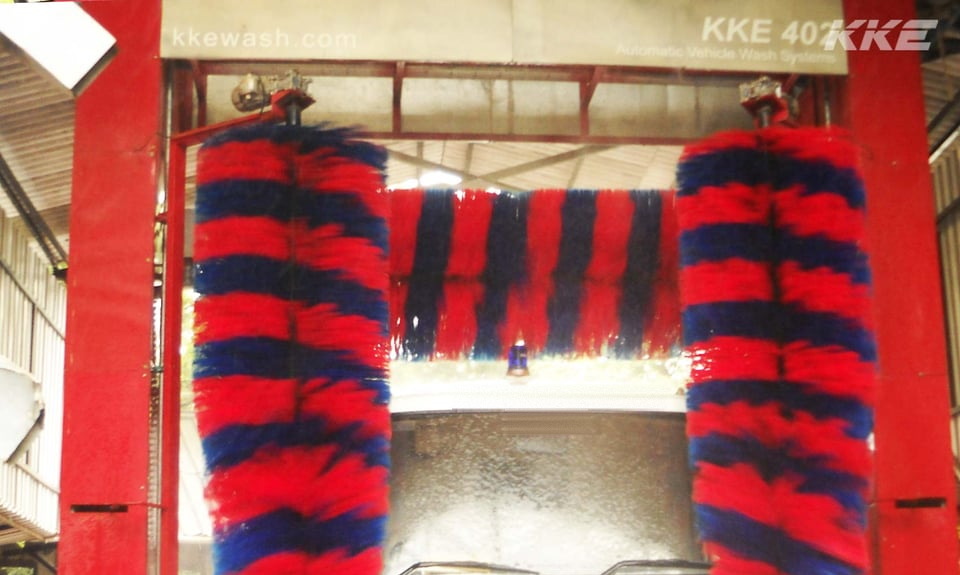 A KKE 403 bus and truck wash seen head-on from inside the bay: a red gantry with a banner reading KKE 403 Automatic Vehicle Wash Systems, a tall red and blue vertical brush hanging either side of the opening and the roof brush turning across the top of the vehicle