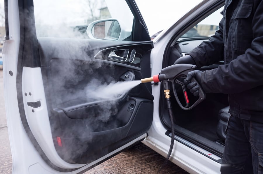 A gloved hand steam cleaning the inside of an open car door, a jet of steam flaring across the leather door card, the armrest, the pull cup and the window switch pack