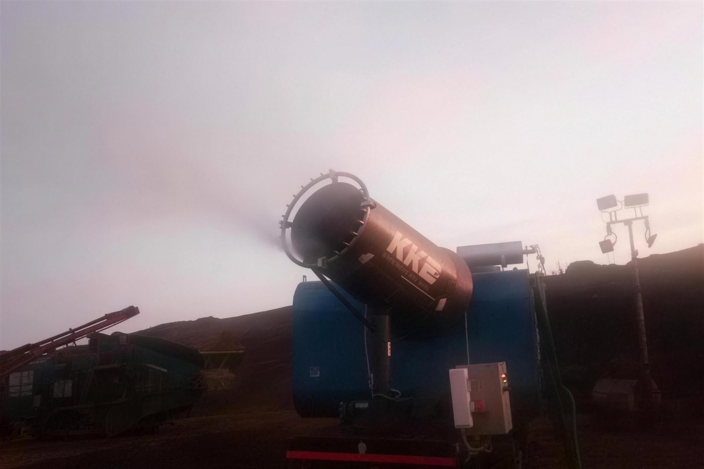 KKE MistBeam fog cannon throwing mist after sundown with site floodlights and a mobile crusher behind it