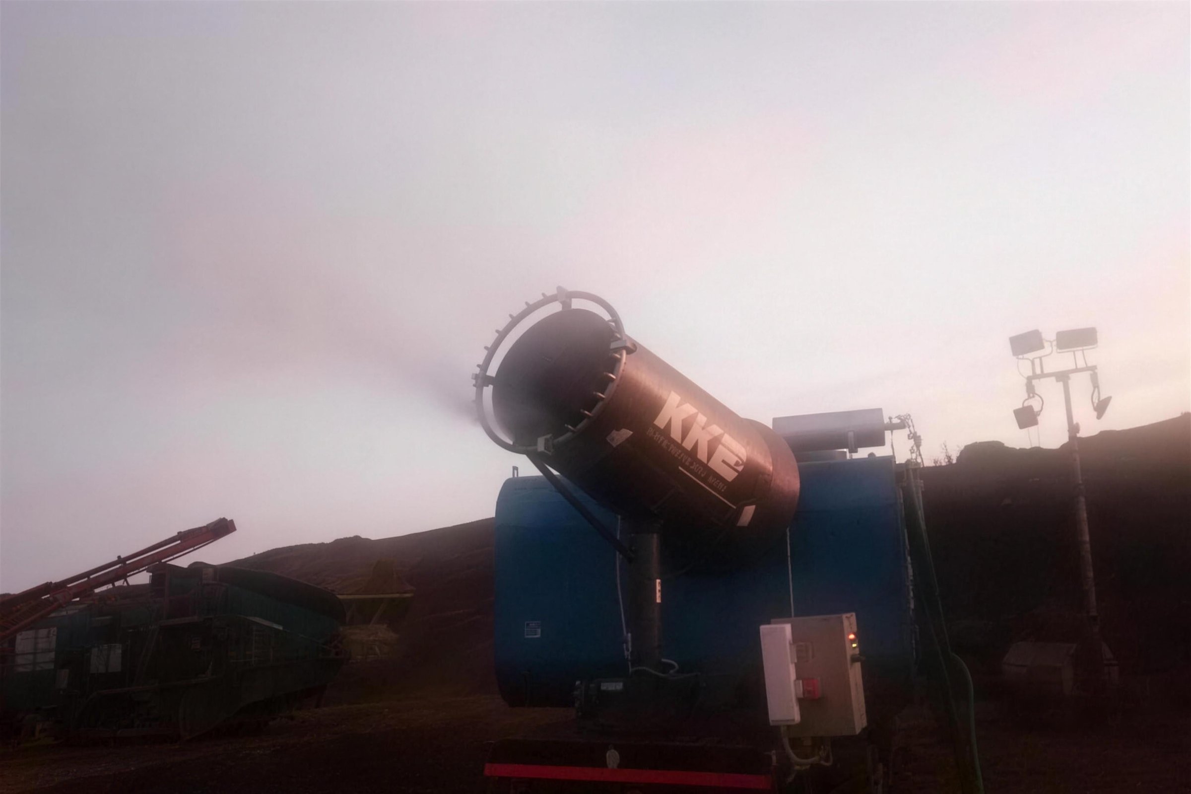 A KKE MistBeam fog cannon throwing mist after sundown with site floodlights and a mobile crusher behind it