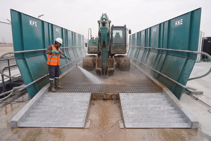 An operator pressure-washing a tracked excavator on a KKE portable wash rack, the splash guard walls catching the spray