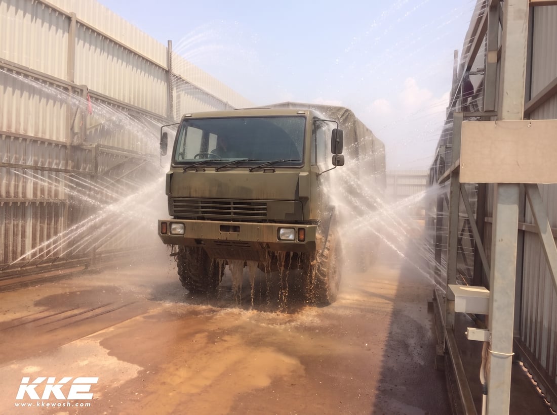 High-pressure jets from the nozzle banks on both walls of a KKE wash lane blasting mud off an olive military truck as it drives through, the under-chassis spray thrown up around its wheels