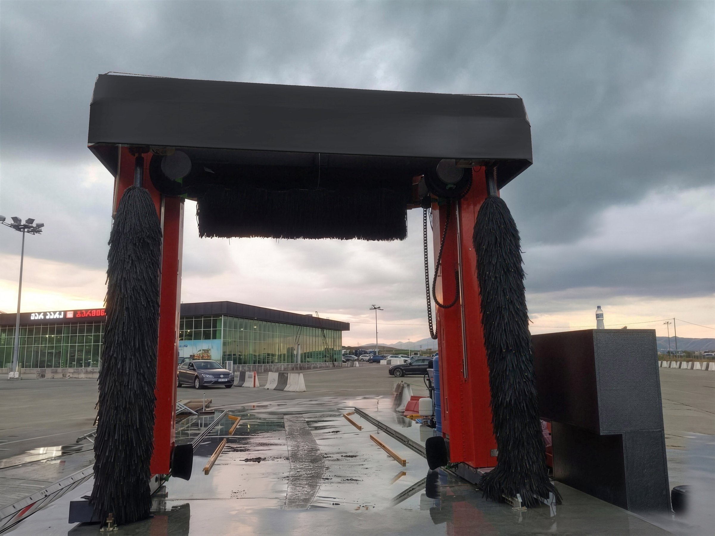 A red KKE X1.1 standing over its wash bay at dusk, the three brushes hanging under the canopy and the forecourt lit behind it