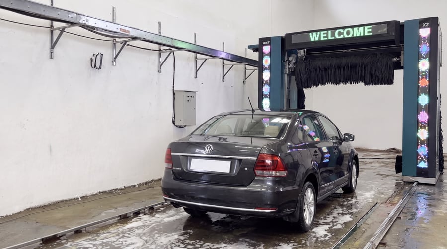 A dark saloon car standing clean and gleaming on the rails at the end of its wash, the KKE X2 gantry parked behind it and the display reading WELCOME