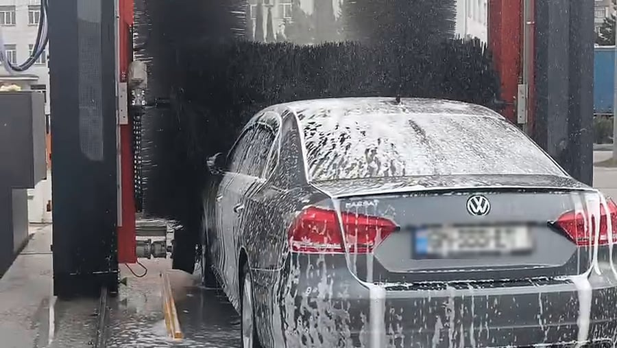 A saloon car covered from roof to sill in white wash foam inside a KKE X1.1 bay in Tbilisi, the brushes parked clear above it