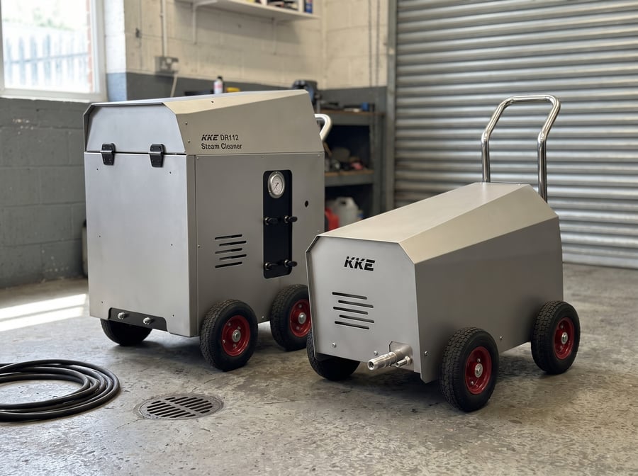 A KKE DR112 steam cleaner and a KKE high pressure cleaner parked together beside the wall of a wash bay, on a concrete floor with a floor drain and a coiled hose