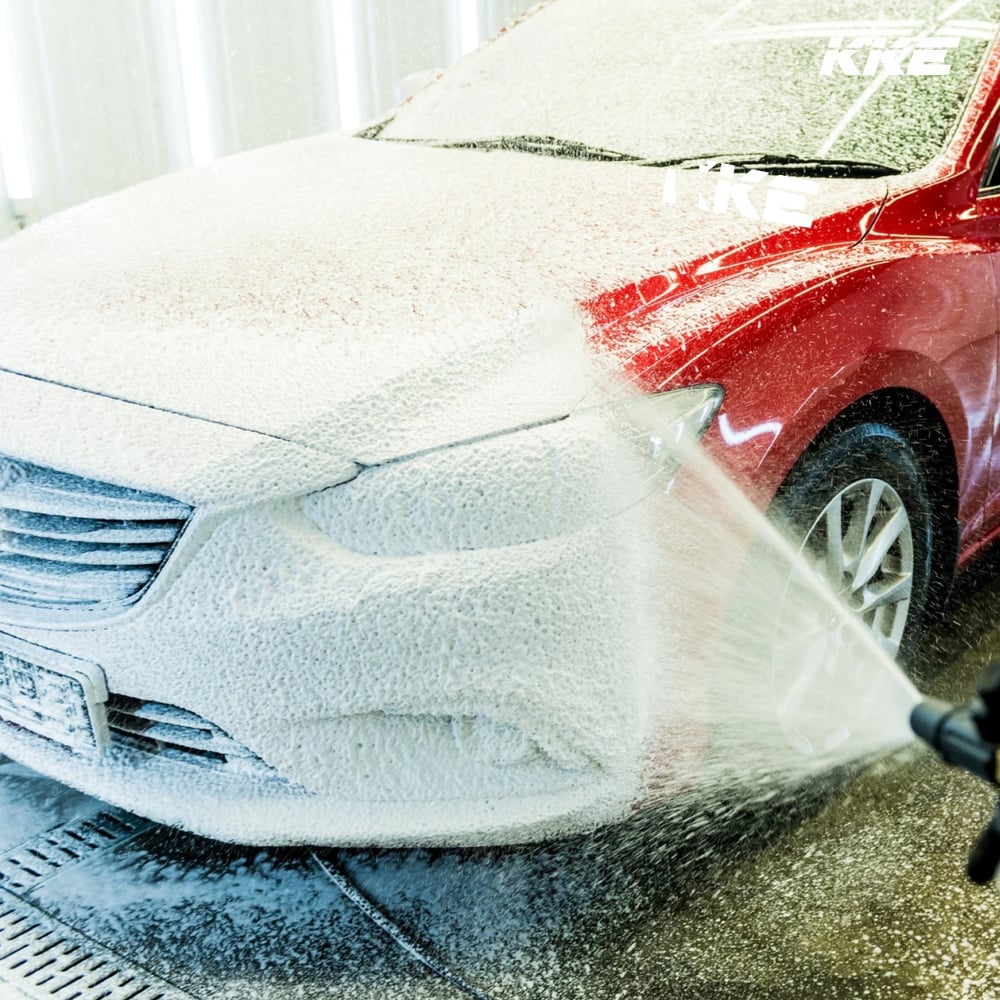 A red car covered in thick KKE snow foam being rinsed with a pressure lance