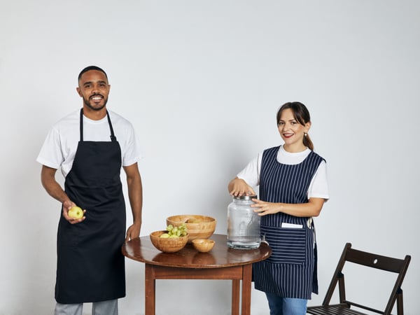 Cozinheiro vestido com um avental preto junto de uma senhora vestida com uma bata avental de cor azul marinho com riscas brancas para serem usadas como peças de fardas de trabalho fabricadas pela Unifardas 