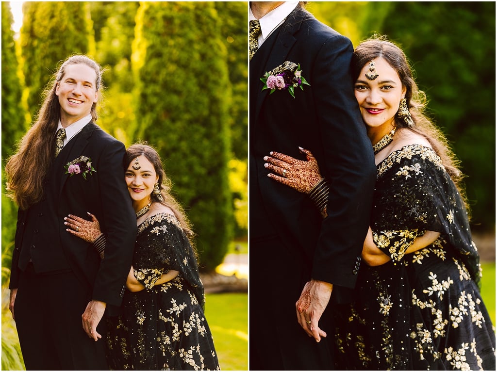 Smiling couple outdoors: a man in a black suit with boutonniere and a woman in a gold-embroidered black outfit; she rests her head on his arm, with henna on her hands.