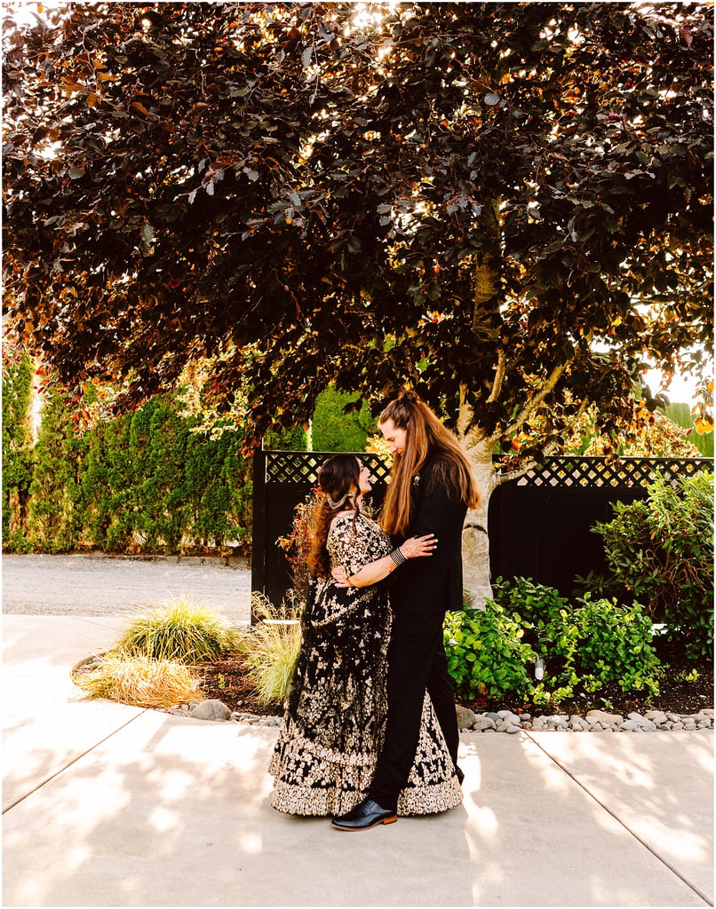 Woman in a black formal dress hugs a girl in a gold-embroidered gown outdoors under a large tree.