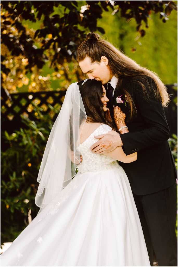 Bride and groom share a kiss/embrace outdoors, bride in a lace wedding dress with veil and henna on her hands, groom in a dark suit with boutonniere, green background.