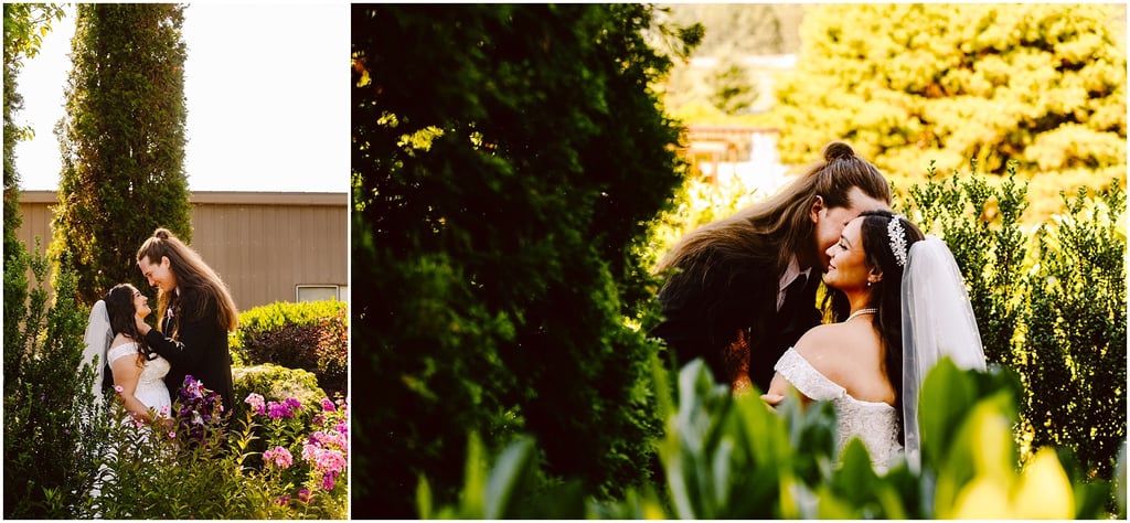 Bride and groom embrace in a sunny garden with pink flowers and tall hedges in the background.",