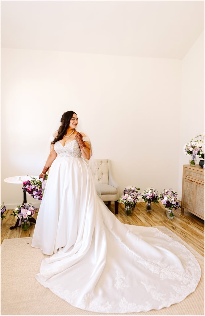 Bride in a white wedding gown with a long train and veil, standing in a bright room with flowers around her.