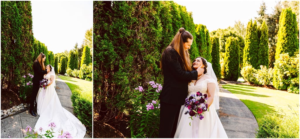 Bride in white gown and veil holds purple bouquet as partner in black suit tenderly holds her face outdoors on a garden path.