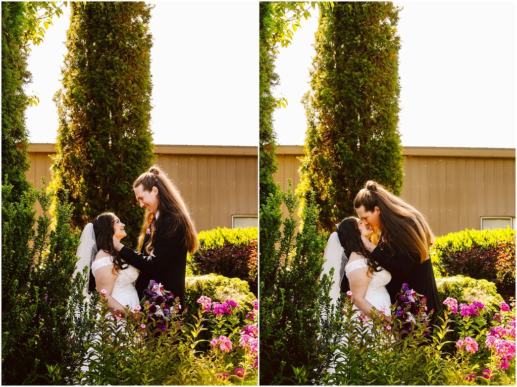 Bride in a lace wedding dress with veil and groom in a dark suit share a kiss in a sunlit garden, with pink flowers and tall shrubs framing them.