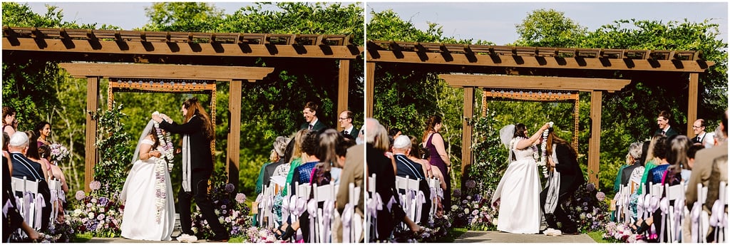 Outdoor wedding ceremony under a wooden arbor with a bride in a white dress and guests seated along the aisle, flowers lining the aisle and decorations in purple and white.
