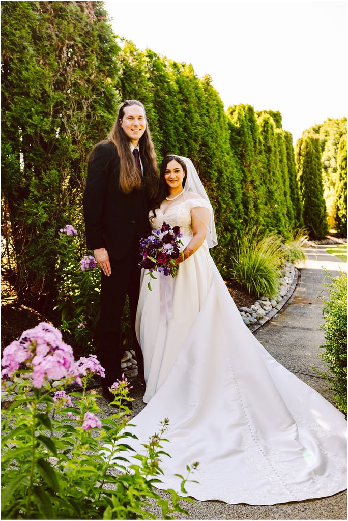 Bride in a white wedding gown and veil holding a purple bouquet stands next to a man in a black suit outdoors along a hedge-lined path, smiling together.