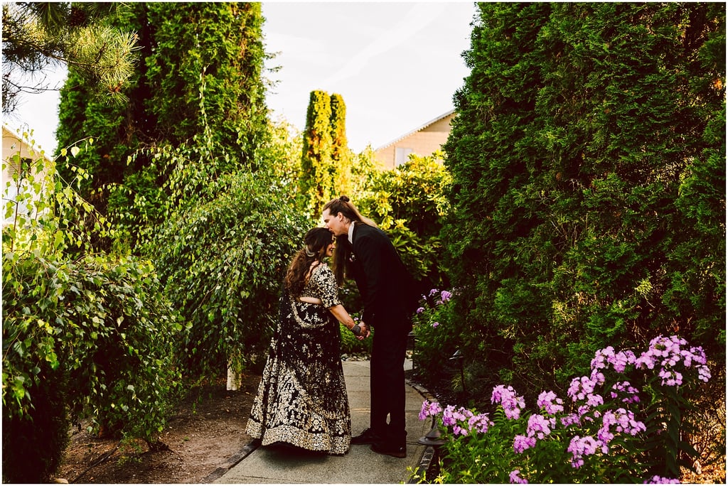 A couple dressed in formal attire holds hands in a sunlit garden path, surrounded by lush green hedges and pink flowers.