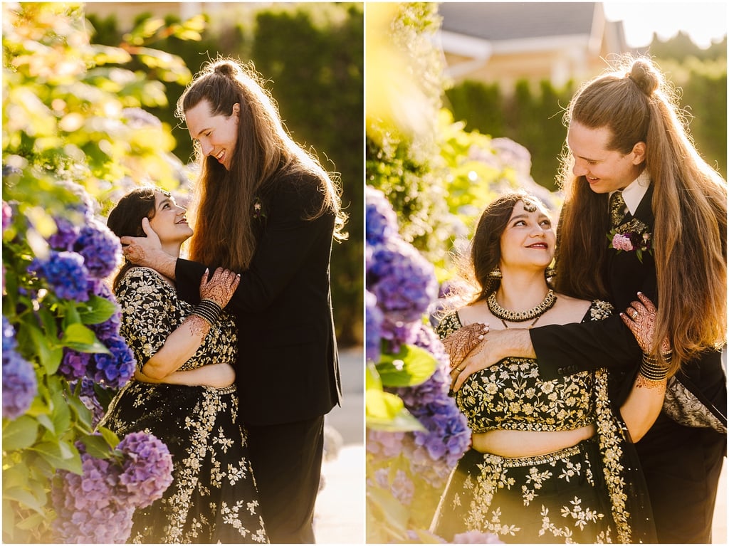 Couple embracing in a sunlit garden among purple hydrangeas; woman in a black and gold embroidery outfit, man in a black suit with long hair tied back.